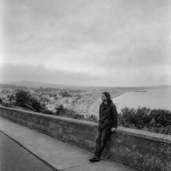 Black-and-white photograph of Nevan Lochlann leaning against a stone wall overlooking Bray Bay and the coastline of County Wicklow, Ireland beneath a cloudy sky